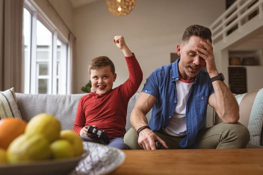 Caucasian boy celebrating while playing video games with his father sitting on the couch at home. gaming and entertainment concept