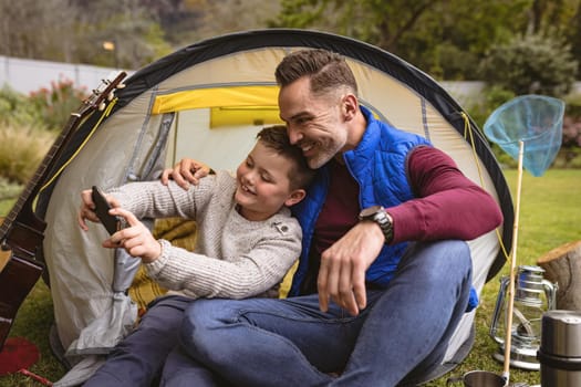 Caucasian father and son taking a selfie while sitting in a tent in the garden. fatherhood and love concept