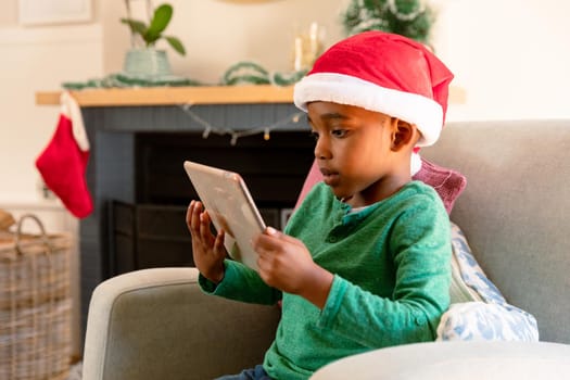 African american boy wearing santa hat and using tablet at christmas time. christmas, festivity and communication technology.