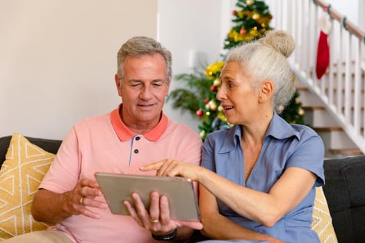 Happy caucasian senior couple sitting on sofa and using tablet at christmas time. healthy retirement lifestyle at home.
