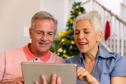Happy caucasian senior couple sitting on sofa and using tablet at christmas time. healthy retirement lifestyle at home.