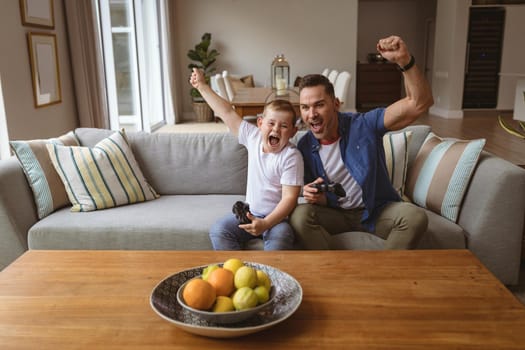 Caucasian father and son celebrating while playing video games sitting on the couch at home. gaming and entertainment concept