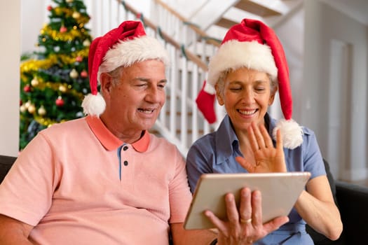 Happy caucasian senior couple wearing santa hat, using tablet, having video call at christmas time. christmas, festivity and communication technology.