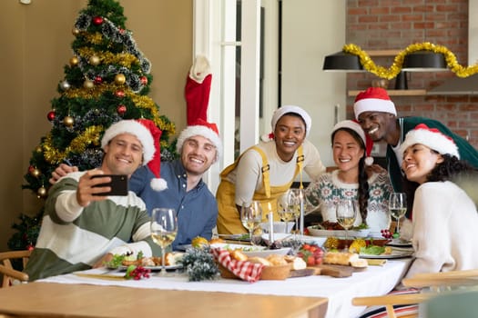 Group of happy diverse female and male friends in santa hats , celebrating christmas, taking selfie. christmas festivities, celebrating at home with friends.