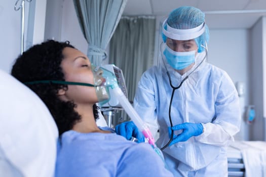 Caucasian doctor in ppe suit examining with stethoscope female patient with oxygen ventilator. medical professional at work during coronavirus covid 19 pandemic.