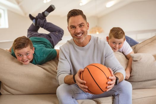 Portrait of caucasian father and two sons with basketball smiling at home. sports and entertainment concept