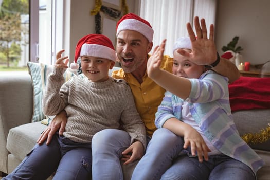 Portrait of caucasian father and two sons waving while sitting on couch at home during christmas. christmas festivity and celebration concept
