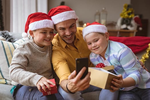 Caucasian father and two sons opening gift box during video call on smartphone during christmas. social distancing during covid 19 pandemic at christmas time