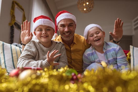 Portrait of caucasian father and two sons smiling while sitting on couch at home during christmas. christmas festivity and celebration concept
