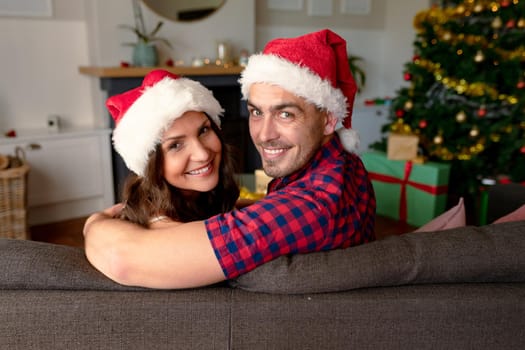 Happy caucasian couple wearing santa hats and siting on sofa at christmas time. christmas, festivity and communication technology.