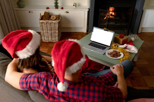 Back view of caucasian couple wearing santa hats, using laptop with copy space at christmas time. christmas, festivity and communication technology.