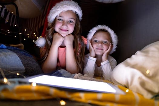 Happy caucasian siblings wearing santa hats and using tablet at christmas time. childhood, chiristmas, festivity and tradition at home.