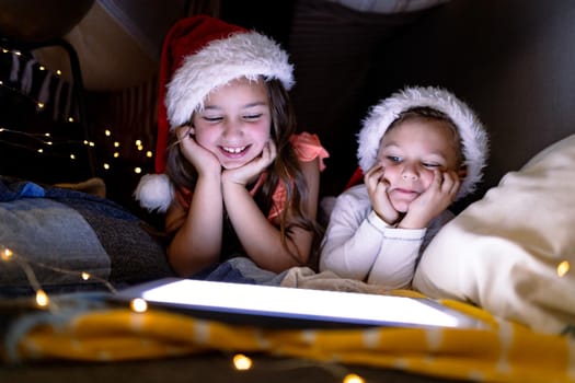 Happy caucasian siblings wearing santa hats and using tablet at christmas time. childhood, chiristmas, festivity and tradition at home.