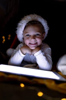 Happy caucasian boy wearing santa hat, using tablet, looking at camera at christmas time. childhood, chiristmas, festivity and tradition at home.