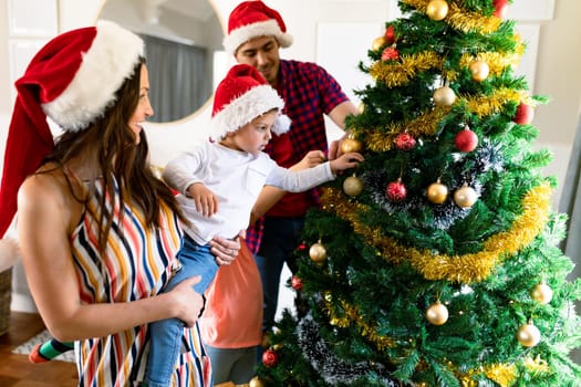 Happy caucasian parents and son wearing santa hats, decorating christmas tree. family christmas time and festivity together at home.