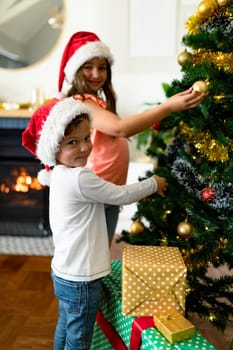 Happy caucasian sibling wearing santa hats, decorating christmas tree. family christmas time and festivity together at home.