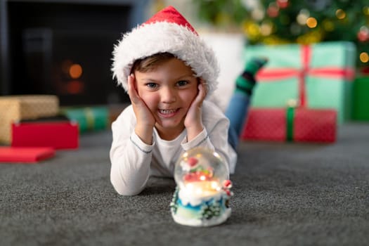 Happpy caucasian boy wearing santa hat, looking at snow globe at christmas time. childhood, chiristmas, festivity and tradition at home.
