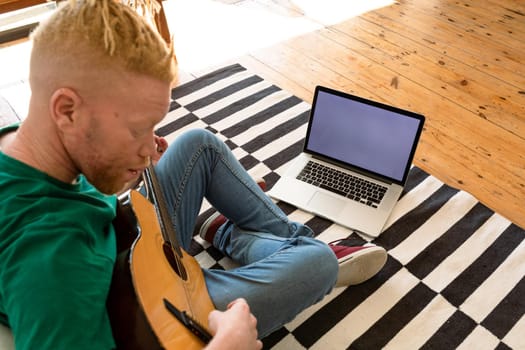 Albino african american man in the living room playing guitar and using laptop. leisure time using technology, relaxing at home.