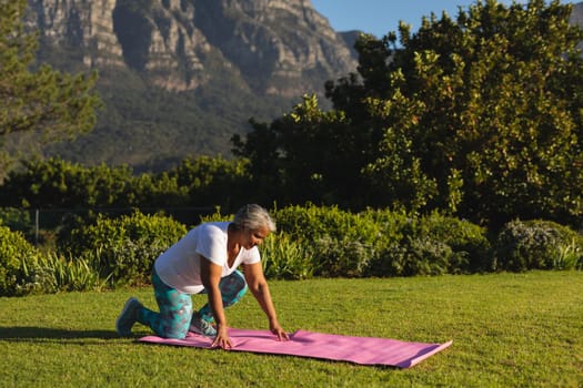 Senior african american woman kneeling on yoga mat on grass in stunning countryside. retirement and active senior lifestyle concept.