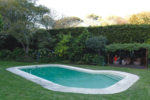 Happy senior caucasian woman sitting on chair, reading book next to swimming pool in garden. retirement lifestyle, spending time alone at home.