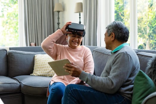 Two happy diverse senior couple wearing vr headset and table and having fun. retirement lifestyle relaxing at home with technology.
