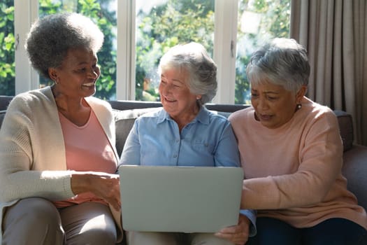 Three happy diverse senior woman sitting on sofa and using laptop. retirement lifestyle relaxing at home with technology.