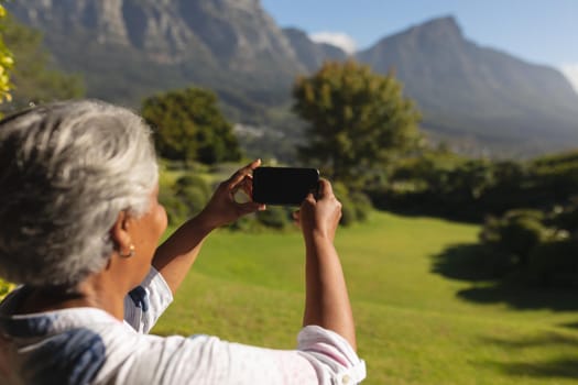 Senior african american woman taking photographs on smartphone of stunning view in sunny garden. retreat, retirement and happy senior lifestyle concept.
