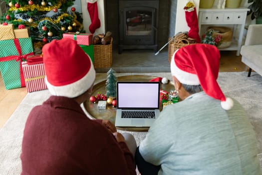 Two diverse senior female friends wearing santa hats using laptop with copy space at christmas time. christmas, festivity and communication technology.