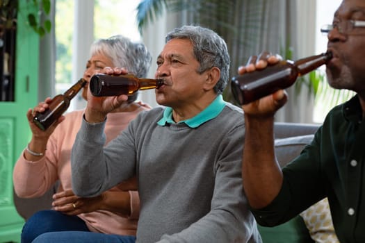 Two diverse senior couple and their african american male friend watching tv and drinking beer. retirement lifestyle relaxing at home with technology.