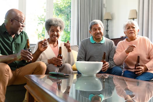 Two diverse senior couples sitting on sofa with beer and having fun. retirement lifestyle relaxing at home with technology.