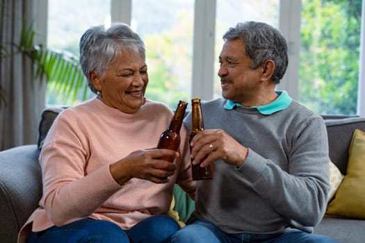 Happy diverse senior couple sitting on sofa and drinking beer. retirement lifestyle and relaxing at home.