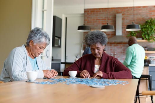 Two diverse female friends sitting in kitchen with coffee and doing puzzles. socialising with friends at home.