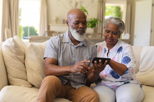 Senior african american couple sitting on sofa using smartphone. retreat, retirement and happy senior lifestyle concept.