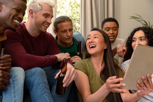 Group of happy diverse female and male friends drinking beer together and using tablet. socialising with friends at home.