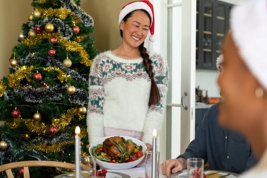 Happy asian woman in santa hat holding dish, celebrating christmas with friends at home. christmas festivities, celebrating at home with friends.