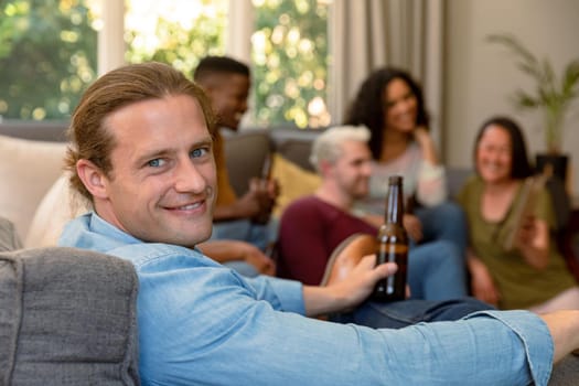 Happy caucasian man holding beer bottle having fun with diverse group of female and male friends. socialising with friends at home.
