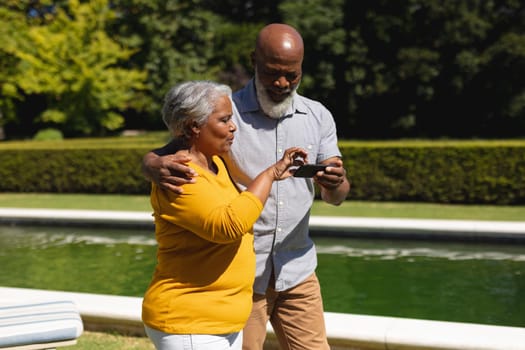Senior african american couple spending time in sunny garden together using smartphone and smiling. retreat, retirement and happy senior lifestyle concept.
