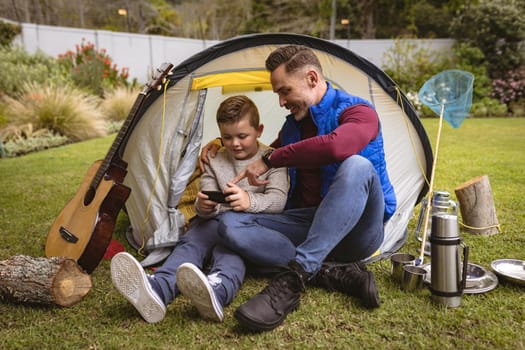 Caucasian father and son using smartphone while sitting in a tent in the garden. fatherhood and love concept