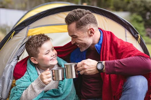 Caucasian father and son smiling while toasting their coffee cups sitting in a tent in the garden. fatherhood and love concept