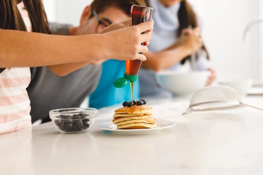 Asian family preparing breakfast in kitchen,father watching daughter put syrup on pancakes. family enjoying preparing meal together at home.