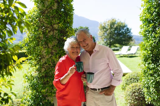 Portrait of senior caucasian couple smiling and holding mugs in sunny garden. retreat, retirement and happy senior lifestyle concept.