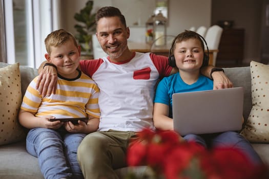 Portrait of caucasian father and two sons with laptop and digital tablet smiling at home. fatherhood and love concept