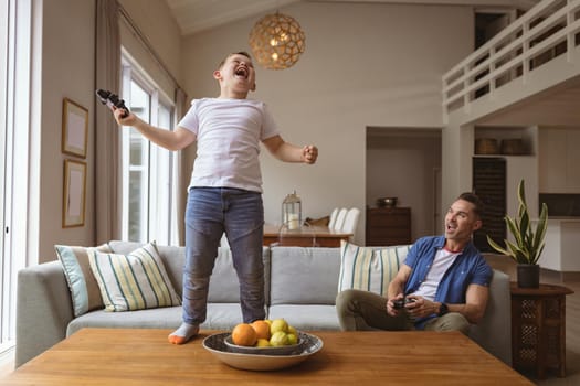 Caucasian boy celebrating standing on the table while playing video games with his father at home. gaming and entertainment concept
