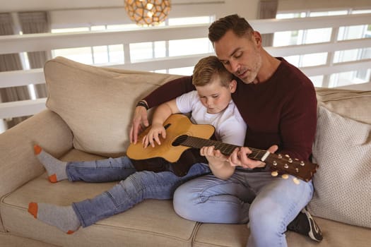 Caucasian father teaching his son to play guitar sitting on the couch at home. childhood and hobby concept