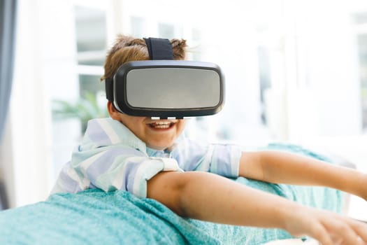 Caucasian boy sitting on couch and using vr headset in living room. childhood leisure time, fun and discovery at at home using technology.