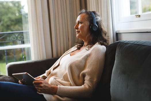 Happy senior caucasian woman in living room sitting on sofa, wearing headphones, using tablet. retirement lifestyle, spending time alone at home with technology.