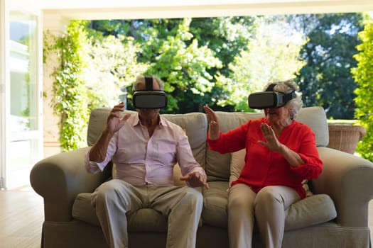 Senior caucasian couple sitting on sofa together wearing vr headset touching virtual screen. retreat, retirement and happy senior lifestyle concept.