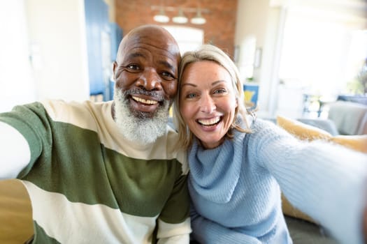 Happy senior diverse couple in living room sitting on sofa, making video call. retirement lifestyle, at home with technology.