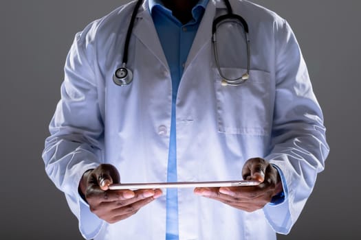 Mid section of african american male doctor holding digital tablet against grey background. healthcare and medical professionalism concept