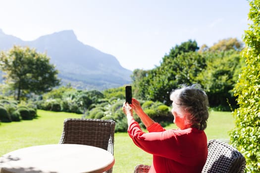 Senior caucasian woman sitting at table using smartphone in sunny garden. retreat, retirement and happy senior lifestyle concept.